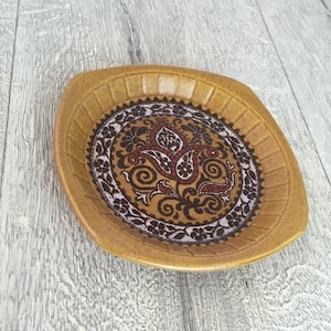 May include: A vintage, mustard-colored ceramic dish with a decorative paisley design in brown, red, and white. The dish has a square shape with rounded corners and a textured rim. The central design features floral and paisley motifs.