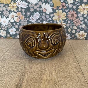 May include: A brown ceramic bowl with a face design. The bowl has a wide opening and a rounded shape. The face features large, spiral eyes, a small nose, and a wide, smiling mouth. The bowl is sitting on a wooden surface.
