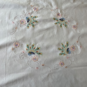 May include: Cream-colored fabric with embroidered peacock and floral designs. The embroidery features blue, green, yellow, and pink thread, creating a decorative pattern. The design is arranged in a circular pattern.