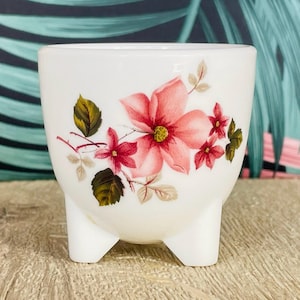 May include: A white ceramic planter with three small feet. The planter is decorated with a floral design featuring pink flowers, green leaves, and delicate stems. The design is centered on the front of the planter.