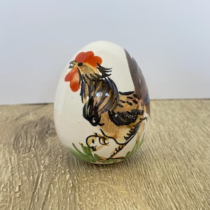 May include: A decorative ceramic egg with a hand-painted rooster design. The rooster has a red comb and wattle, with brown and black feathers. Green grass is painted at the base of the egg, set on a wooden surface.