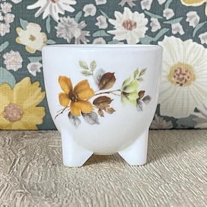 Könnte beinhalten: Kleiner, weißer Keramik-Blumentopf mit drei kurzen Beinen. Der Topf ist mit einem Blumendesign mit gelben und braunen Blumen und grünen und grauen Blättern verziert. Der Hintergrund ist ein Blumenmuster.