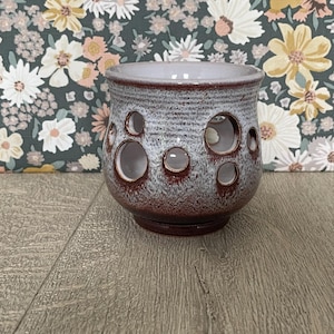 May include: A ceramic candle holder with a rounded shape, featuring a textured light grey upper section and a deep reddish-brown base. The exterior is decorated with several circular cutouts of varying sizes, revealing the smooth white interior.