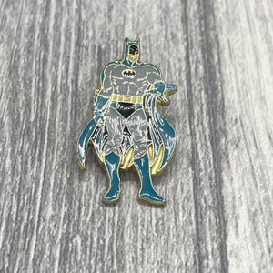 May include: A metal enamel pin featuring a standing figure of Batman. The character is depicted in a grey suit with a black bat emblem on the chest, a black utility belt, and dark teal gloves, boots, and a flowing cape with gold-toned trim.