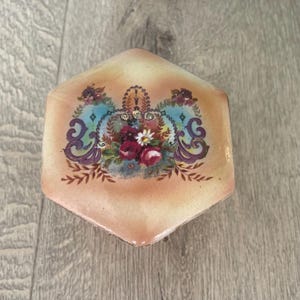 May include: A hexagonal lidded box with a glossy peach-coloured finish. The lid features a central floral arrangement of red and white flowers, surrounded by intricate blue and purple scrollwork patterns and delicate decorative filigree details.