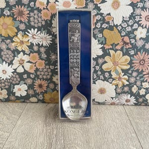 May include: A silver-colored souvenir spoon with intricate engraved designs and the text "KONGEN Norwegian pewter." The spoon is displayed in a blue box against a floral patterned background.