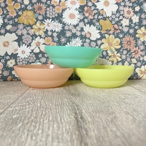 Könnte beinhalten: Drei pastellfarbene Schalen in Pfirsich, Mintgrün und Gelb sind gestapelt. Die Schalen haben eine abgerundete Form und eine glänzende Oberfläche. Der Hintergrund zeigt ein Blumenmuster. Die Schalen sind aus Glas oder Keramik.