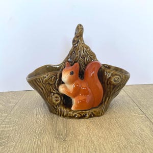May include: A ceramic basket-shaped planter with a brown tree bark design. A glossy orange squirrel figurine is nestled inside. The handle is a twisted tree branch. The planter is on a light wood surface.