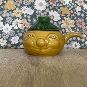 May include: A yellow ceramic pot with a smiling face, a round nose, and a handle on the right side. It features a green lid with the embossed text PAN SCOUR. The pot has a glossy finish and a rounded body, designed for kitchen utility.