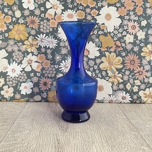 May include: A vibrant cobalt blue glass vase with a flared top and a rounded base. The vase has a smooth, glossy finish and stands on a small, circular foot. The background features a floral pattern with various flowers.