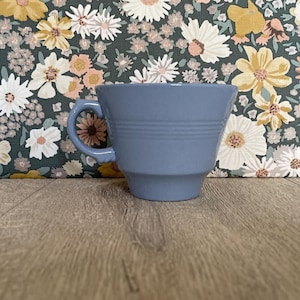 May include: A periwinkle blue ceramic cup featuring a wide, flared rim and a tapered base. The exterior of the cup is decorated with three horizontal embossed rings encircling the middle section, complemented by a curved handle on the left side.