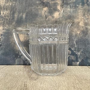 May include: A clear glass creamer pitcher featuring a vertical ribbed pattern on the lower body, a band of diamond-shaped geometric embossing around the center, and a scalloped rim. The pitcher includes a sturdy handle and a small pouring spout.