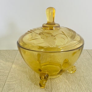 May include: Amber-coloured glass candy dish with a lid and three feet. The lid features an embossed floral design and a handle. The dish is a vintage style and is likely used for storing small items or sweets.