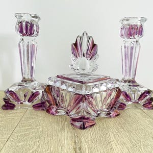 May include: A set of three clear glass decorative items with purple accents. The set includes two matching candlesticks and a square-shaped trinket box with a decorative lid. The glass has a faceted design.