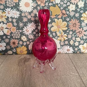 May include: A vibrant, ruby-red glass vase with a bulbous base and slender neck. The vase sits on a clear, three-legged stand. The background features a floral pattern with white, yellow, and pink flowers on a dark green backdrop.