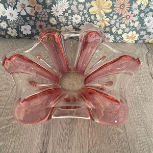 May include: A clear glass bowl with a pink floral design. The bowl has a star-like shape with pink petals radiating from the centre. The bowl is sitting on a wooden surface.