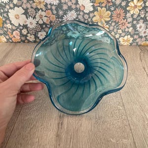 May include: A teal-coloured glass bowl with a distinctive, wavy edge. The interior of the bowl has a spiral pattern emanating from a central hole. The bowl is held by a hand, highlighting its delicate shape and colour.