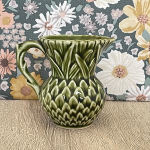 May include: A glossy olive green ceramic pitcher featuring a textured pineapple-inspired design. The body of the pitcher displays a repeating scale pattern, while the upper section is adorned with sculpted leaf motifs and a curved handle.