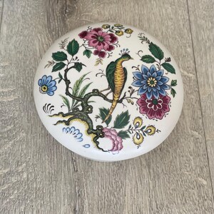 May include: A round white ceramic trinket box lid featuring a colorful hand-painted design of a bird perched on a tree branch, surrounded by various flowers in shades of pink, blue, and yellow with green leaves, all set against a smooth surface.
