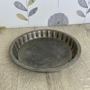 May include: A round, fluted metal pie pan with a dark, aged appearance. The pan has a slightly raised edge and a flat base. The pan is sitting on a wooden surface.