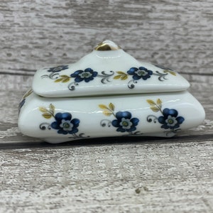 May include: A white porcelain trinket box with a lid, adorned with blue floral patterns and gold accents. The box has a curved, rounded shape and sits on a wooden surface. The floral design includes blue flowers and gold leaves.