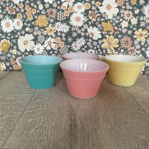 May include: A set of four small, round bowls in solid pastel colors including turquoise, light pink, and yellow. Each bowl features a smooth, tapered design with a subtle horizontal ridge detail near the top rim, arranged in a cluster on a surface.