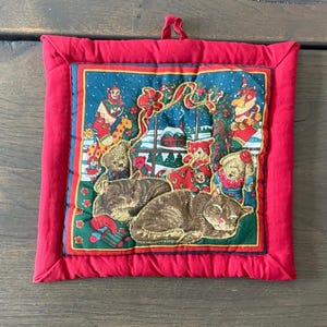 May include: A red quilted Christmas potholder with a square design. The centre features a festive scene with sleeping cats, teddy bears, and snow-covered houses. The border is a solid red.