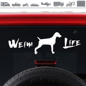 Op de afbeelding: Witte silhouette van een Weimaraner met de woorden "Weim" en "Life" in een gestileerd lettertype op een zwarte achtergrond. De afbeelding is een auto-sticker voor het raam.
