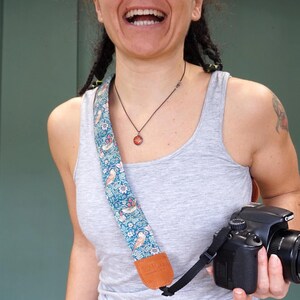 May include: A woman wearing a gray tank top and a floral camera strap is holding a black Canon DSLR camera with a lens.