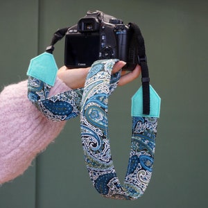 May include: A black camera with a paisley patterned camera strap. The strap has turquoise blue accents.
