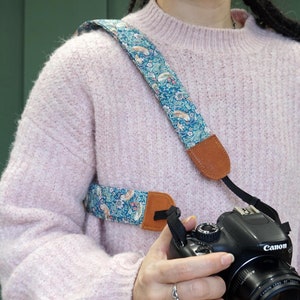 May include: A blue and white floral camera strap with brown leather accents. The strap is worn over a person's shoulder and is attached to a black Canon camera.