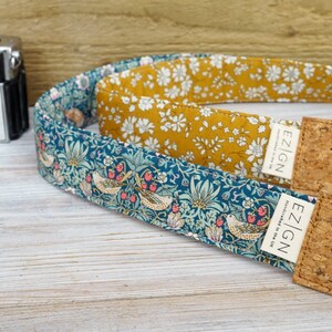 Puede incluir: Dos correas de cámara con extremos de corcho. Una correa es de color turquesa con un patrón floral y pájaros. La otra correa es de color amarillo mostaza con un patrón floral blanco. Ambas correas tienen el texto "EZ|GN" impreso.