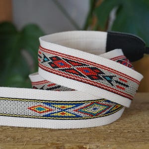 May include: White camera strap with a woven pattern in red, black, blue, and yellow geometric designs. The strap is made of a sturdy material and has a black attachment piece.