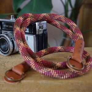 May include: A retro camera with the words "INSTANT LOAD" printed on the top, alongside a colourful rope camera strap. The strap has a burgundy, tan, and gold pattern, with brown leather ends and metal rings.