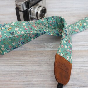 Op de afbeelding: Een groene cameraband met een bloemenpatroon en bruine leren accenten. De band is gemaakt van stof en heeft een verstelbare lengte.