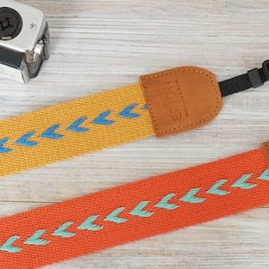 Pode incluir: Duas correias de câmera com comprimento ajustável, uma amarela com padrão de chevron azul e outra laranja com padrão de chevron turquesa. Ambas as correias têm pontas de couro castanho com a palavra "Eizen" gravada.