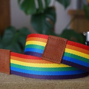 Op de afbeelding: Een regenboogkleurige camera-riem met een bruine leren accent. De riem is gemaakt van geweven stof en heeft een regenboogpatroon.
