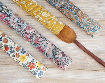 May include: Four colorful camera straps with floral and paisley patterns. One strap is mustard yellow with white flowers, another has a brown leather end piece with the brand name. The straps are on a light wood surface.