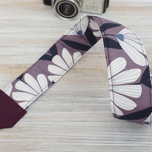 Pode incluir: Uma correia de câmera roxa com um padrão floral branco. A correia tem um detalhe em couro bordô.
