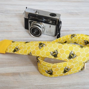 May include: A yellow camera strap with a bee and honeycomb pattern. The strap is made of fabric and has a black strap connector. A vintage silver camera is in the background.
