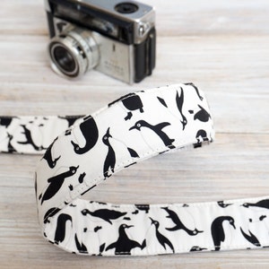 Pode incluir: Uma correia de câmera branca com um padrão de pinguim preto. A correia é feita de tecido e tem uma borda costurada.