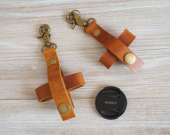 May include: Two brown leather keychains with brass-colored clasps and rivets. The keychains have a cross-shaped design. A black Sony lens cap is also in the image. The keychains are on a light wood surface.