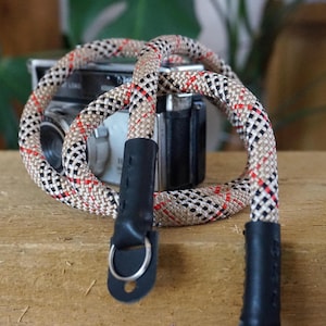 May include: A camera strap with a beige, black, and red rope pattern. The strap is looped around a vintage camera. The ends of the strap have black leather attachments with metal rings.