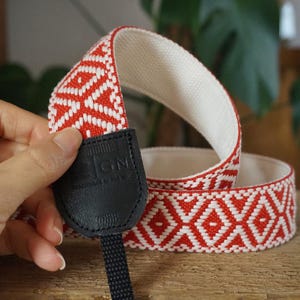 May include: A red and white patterned camera strap with a black leather end piece. The strap features a geometric diamond design. The interior of the strap is white. The black leather piece has the letters "IGN" printed on it.