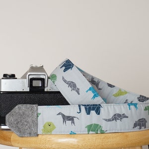 Peut inclure: Un appareil photo gris avec une sangle d'appareil photo à motifs gris et blancs représentant des animaux géométriques. La sangle est en tissu et possède un coussin en feutre gris.