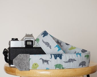 May include: A gray camera with a gray and white patterned camera strap featuring geometric animal designs. The strap is made of fabric and has a gray felt pad.