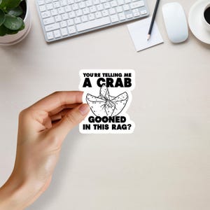 Op de afbeelding: Een witte sticker met zwarte tekst met de tekst "YOU'RE TELLING ME A CRAB GOONED IN THIS RAG?" De sticker heeft een afbeelding van een geknoopte doek. De sticker wordt door een hand vastgehouden.