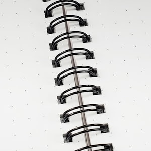 K&ouml;nnte beinhalten: Nahaufnahme einer schwarzen Wire-O-Bindung auf einem wei&szlig;en Notizbuch mit kleinen schwarzen Punkten.