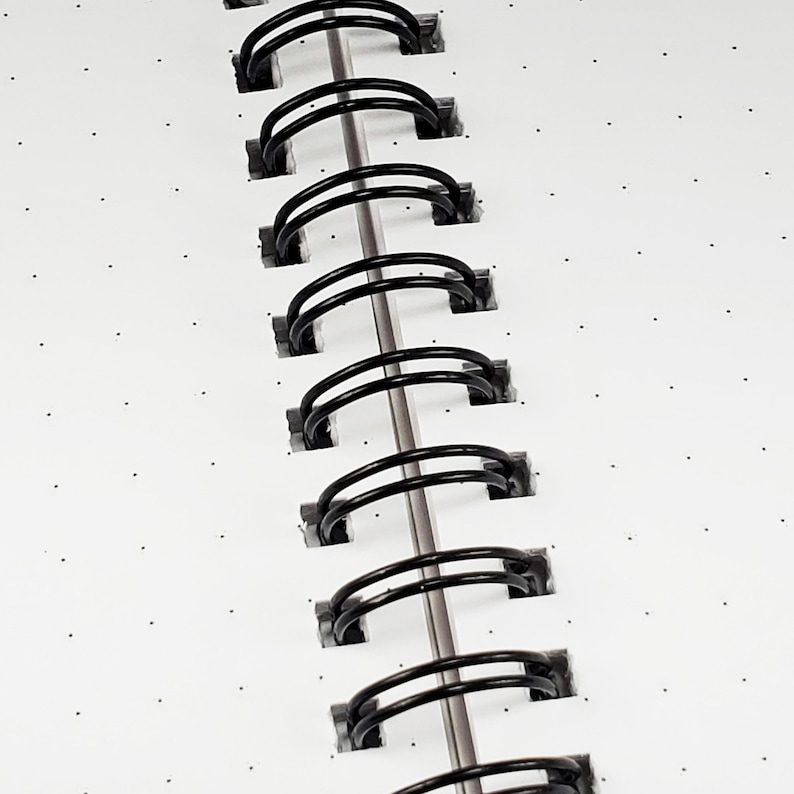 K&ouml;nnte beinhalten: Nahaufnahme einer schwarzen Wire-O-Bindung auf einem wei&szlig;en Notizbuch mit kleinen schwarzen Punkten.