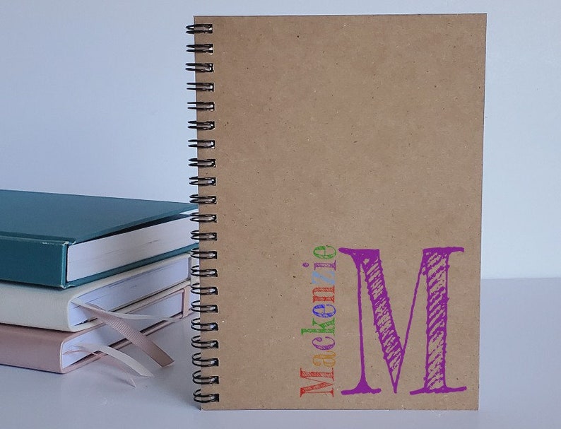 Personalized notebook with brown kraft cover and a colorful name and monogram initial in the lower right side of the cover. These custom notebooks are great for girls or boys, and women. Perfect gift idea for party favors, students and teachers.